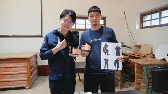 高雄岡山「剛好販売所」店主羅健福（右）左手拿著將赴日展出的環保永續服飾展板，右手拿著岡山青創品牌「俞煙烤肉醬」，與青年局長林楷軒（左）合影。該店結合書店、社區創意與永續理念，曾獲113年競爭型青創補助及111年青創貸款支持，為地方注入新的文化能量。