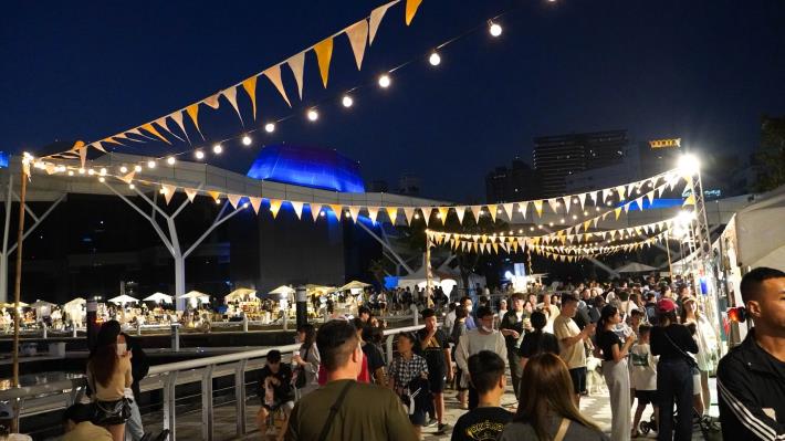 夜幕下的「城市嶼浪」市集燈火通明，民眾漫步愛河灣畔，感受港都悠閒節奏與創意交織的迷人夜色。